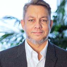 A middle-aged man with short, light brown hair and a trimmed gray beard is wearing a white shirt and a dark checkered blazer. He stands indoors with blurred green plants in the background.