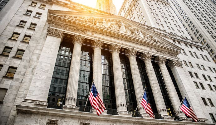 The facade of the New York Stock Exchange building with tall columns, American flags, and sunlight shining from above, surrounded by skyscrapers.