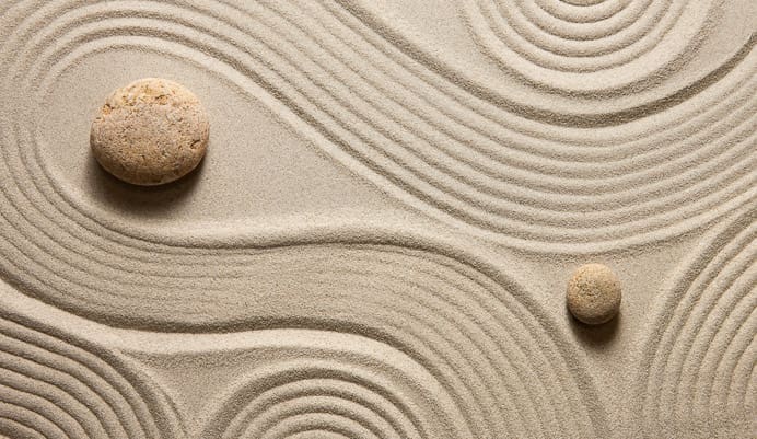 Two smooth stones rest on fine sand with curved, raked lines creating patterns around them, resembling a Zen garden and evoking a sense of calm and tranquility.