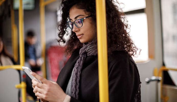A woman with curly hair and glasses stands on a bus, looking at her smartphone. She is wearing a dark coat and scarf, holding onto a yellow pole. Other passengers are blurred in the background.