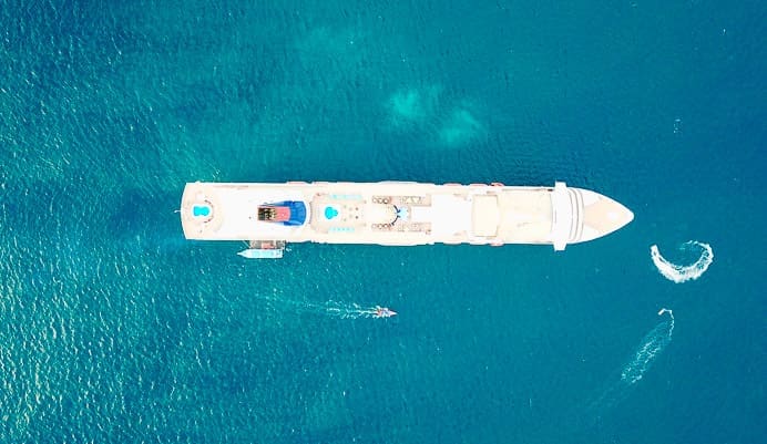 A large white cruise ship is seen from above in turquoise blue water. Smaller boats create gentle wakes nearby, and the ships pool and decks are visible.