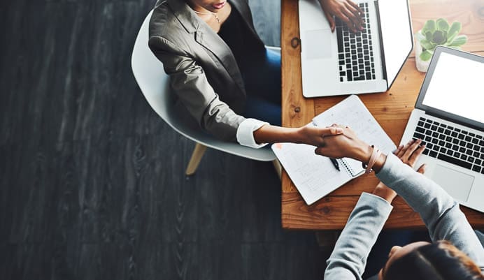 Two people sit at a wooden table with laptops and notebooks, shaking hands in agreement. The scene is viewed from above, suggesting a professional meeting or job interview.