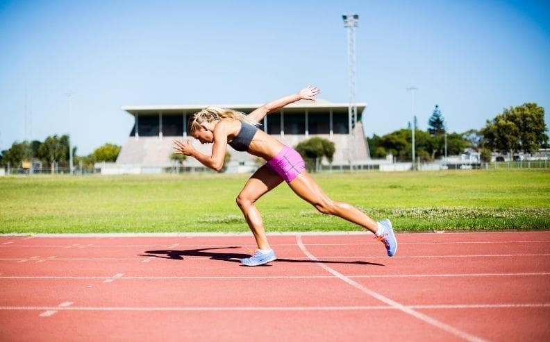 A woman in athletic wear sprints on a track in an outdoor stadium, leaning forward with one knee bent and arms pumping, with green grass and a building visible in the background.