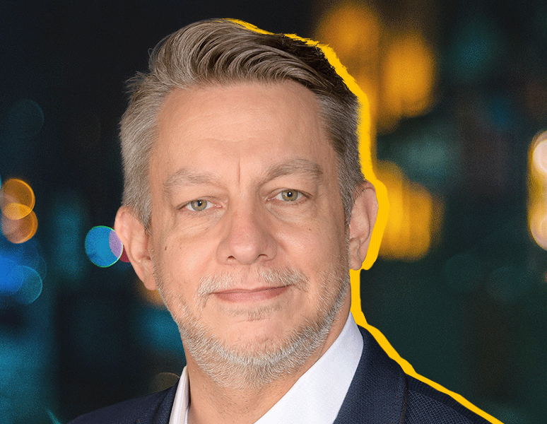 A middle-aged man with short gray hair and a trimmed beard smiles slightly. He is wearing a dark suit and white shirt. The background is blurred with colorful lights, and a yellow outline surrounds his head and shoulders.