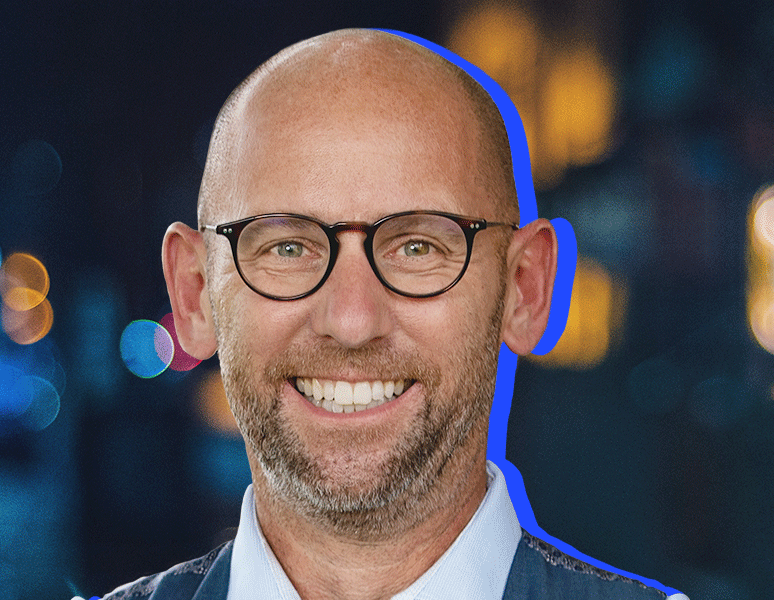 A smiling bald man with a beard and glasses, wearing a collared shirt and jacket, is pictured against a blurred, dark background with colorful lights. A blue outline highlights his profile.
