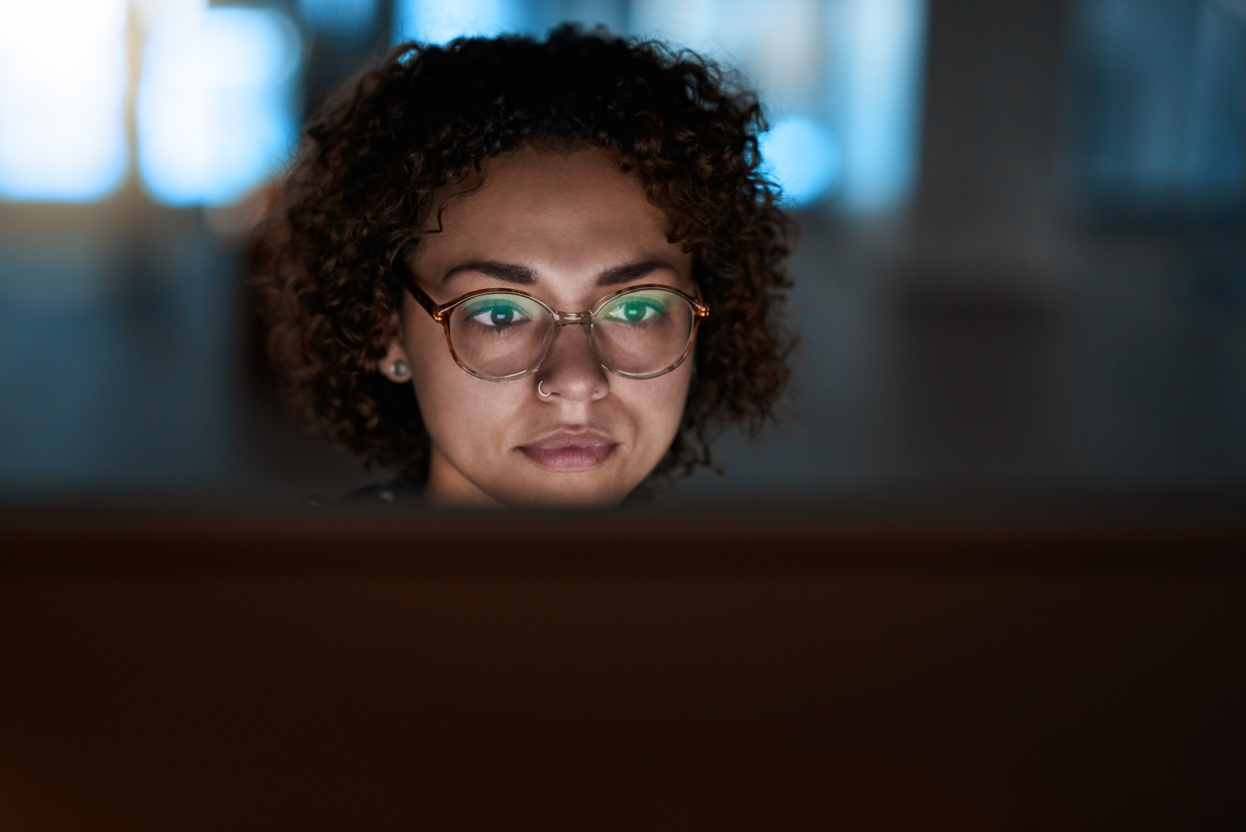 A person with curly hair and glasses is intently looking at a computer screen in a dimly lit room, with the glow of the screen reflecting in their glasses.