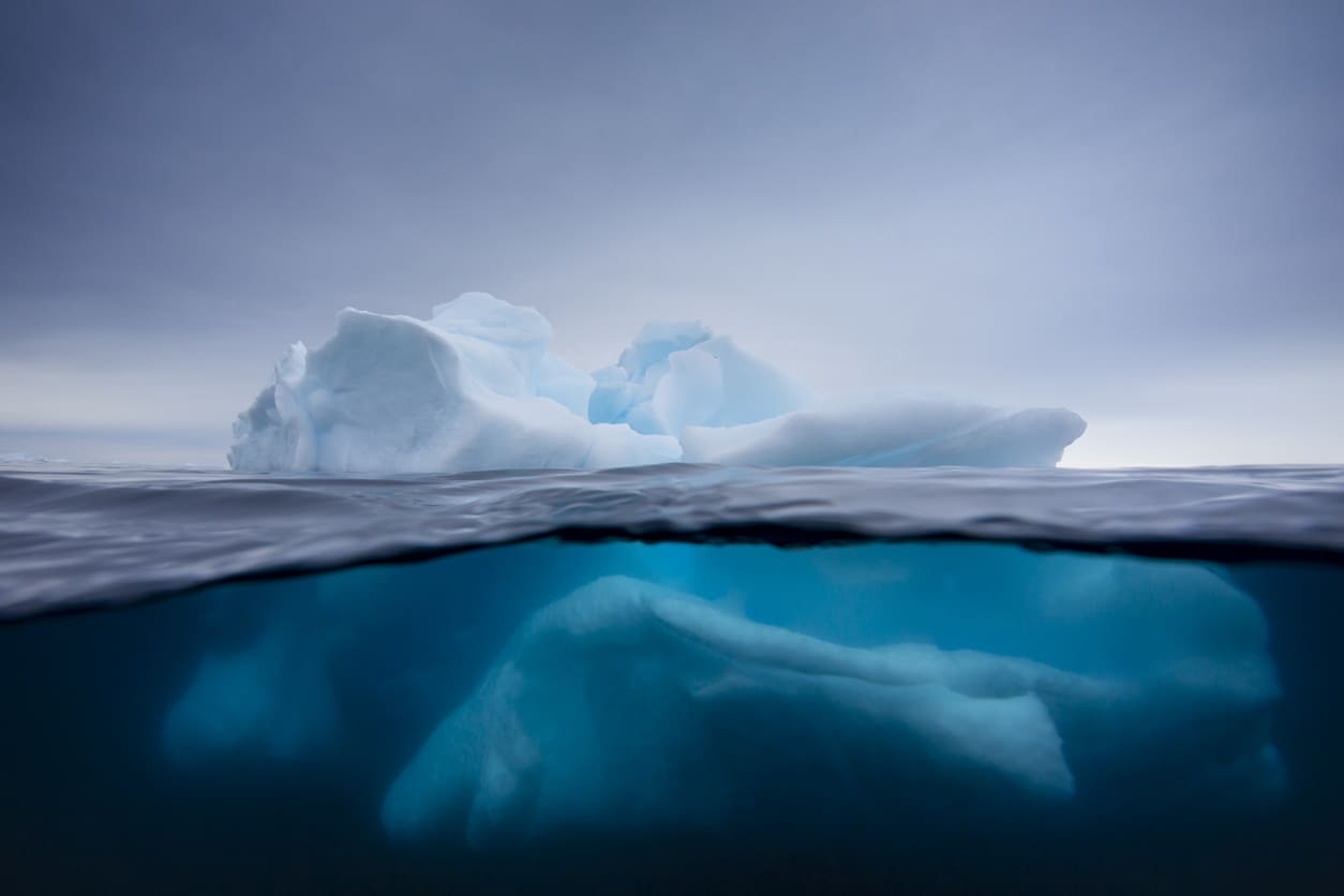 A large iceberg floats in the ocean, with its white tip above the surface and a much larger portion submerged underwater, viewed through clear blue water under a cloudy sky.