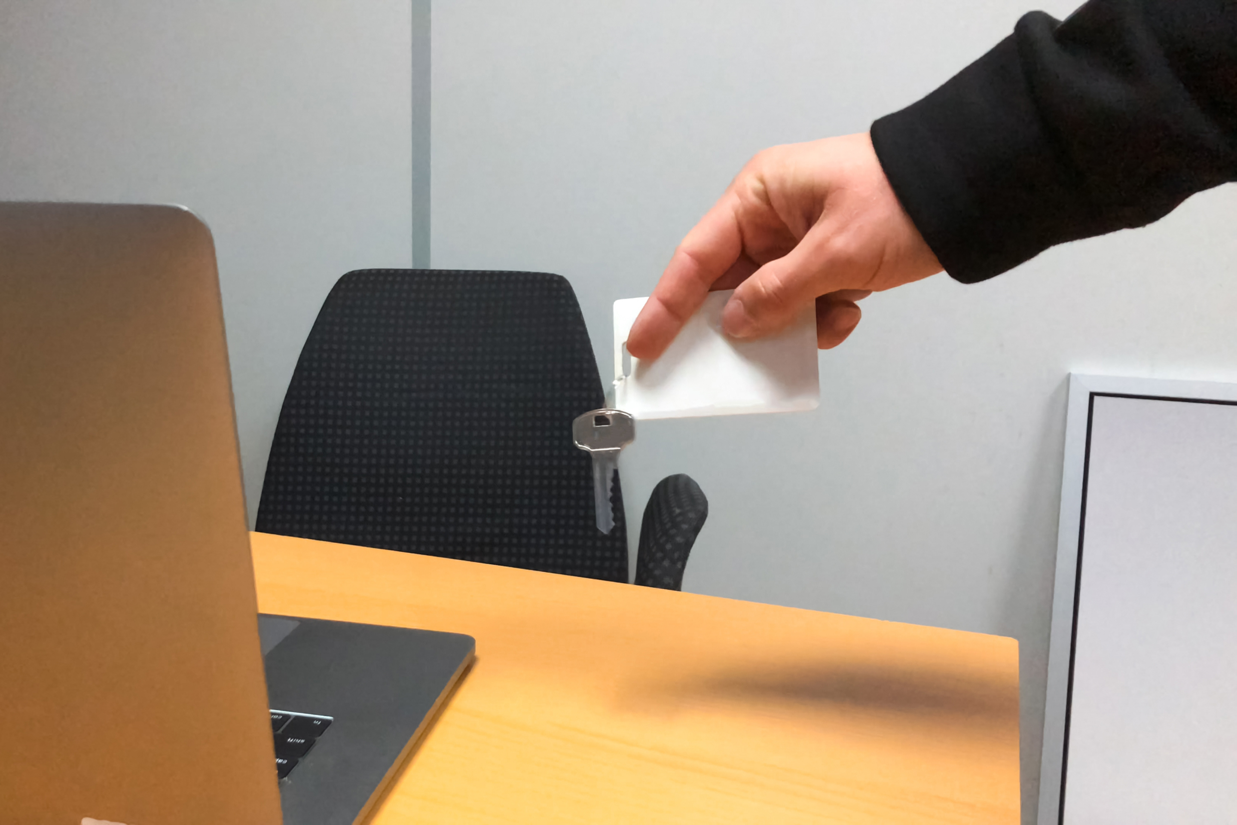 A hand holds a silver key and a white card above a wooden desk with a laptop, an empty black office chair, and a light gray wall in the background.