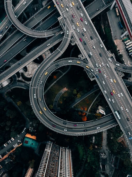 Aerial view of a busy multi-level highway interchange with looping ramps and numerous cars. Green spaces and buildings surround the intricate road network.