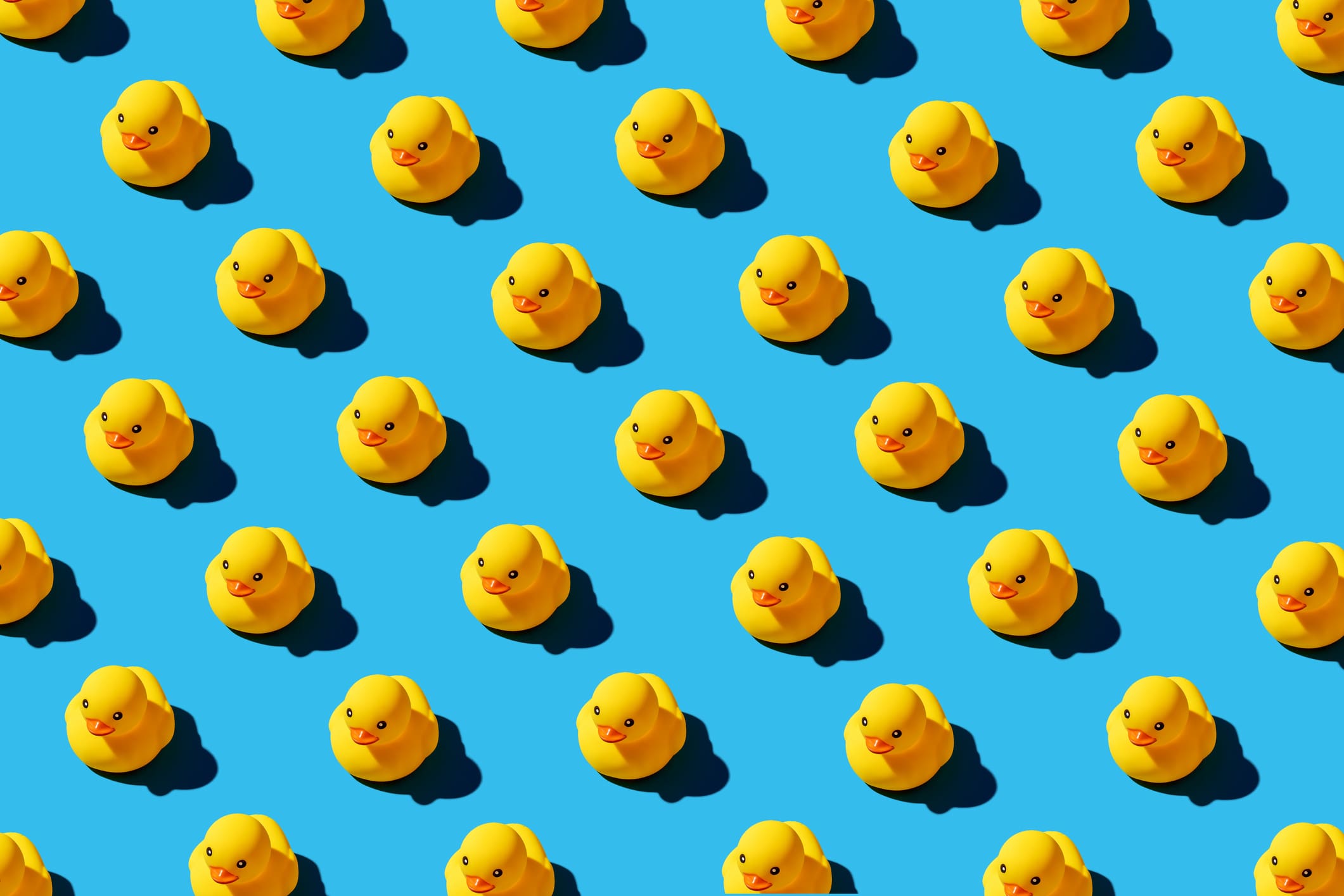 A pattern of yellow rubber ducks arranged in evenly spaced rows on a bright blue background, each casting a shadow to the lower right.