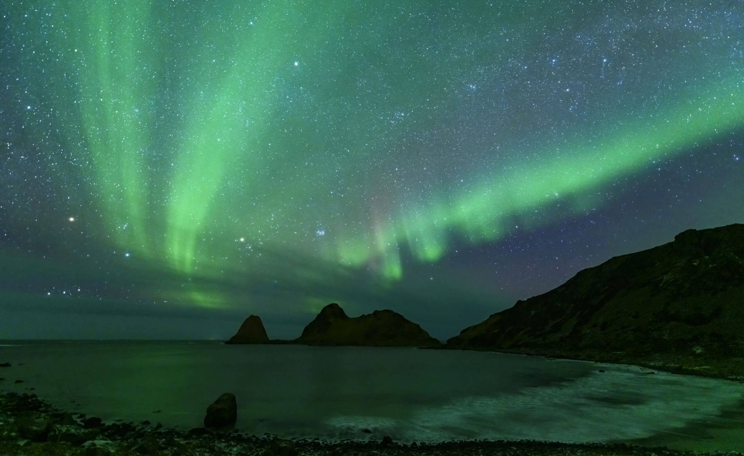 Green aurora borealis lights streak across a starry night sky above a calm sea and rugged, dark silhouetted mountains along a rocky shoreline.