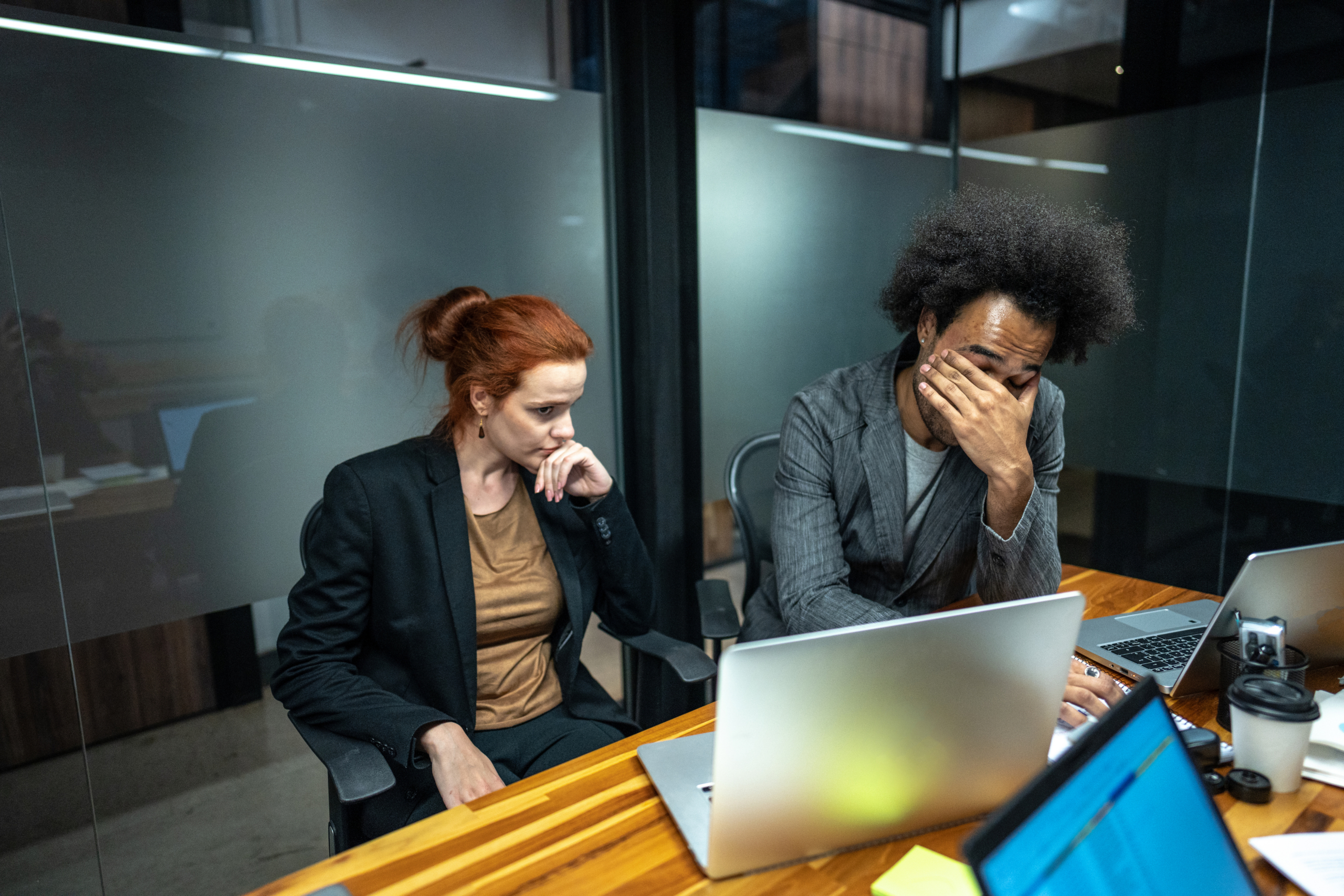 Two people sit at a conference table with laptops, both looking concerned. One person holds their head in their hand, while the other looks at a screen thoughtfully. The office setting appears serious and tense.