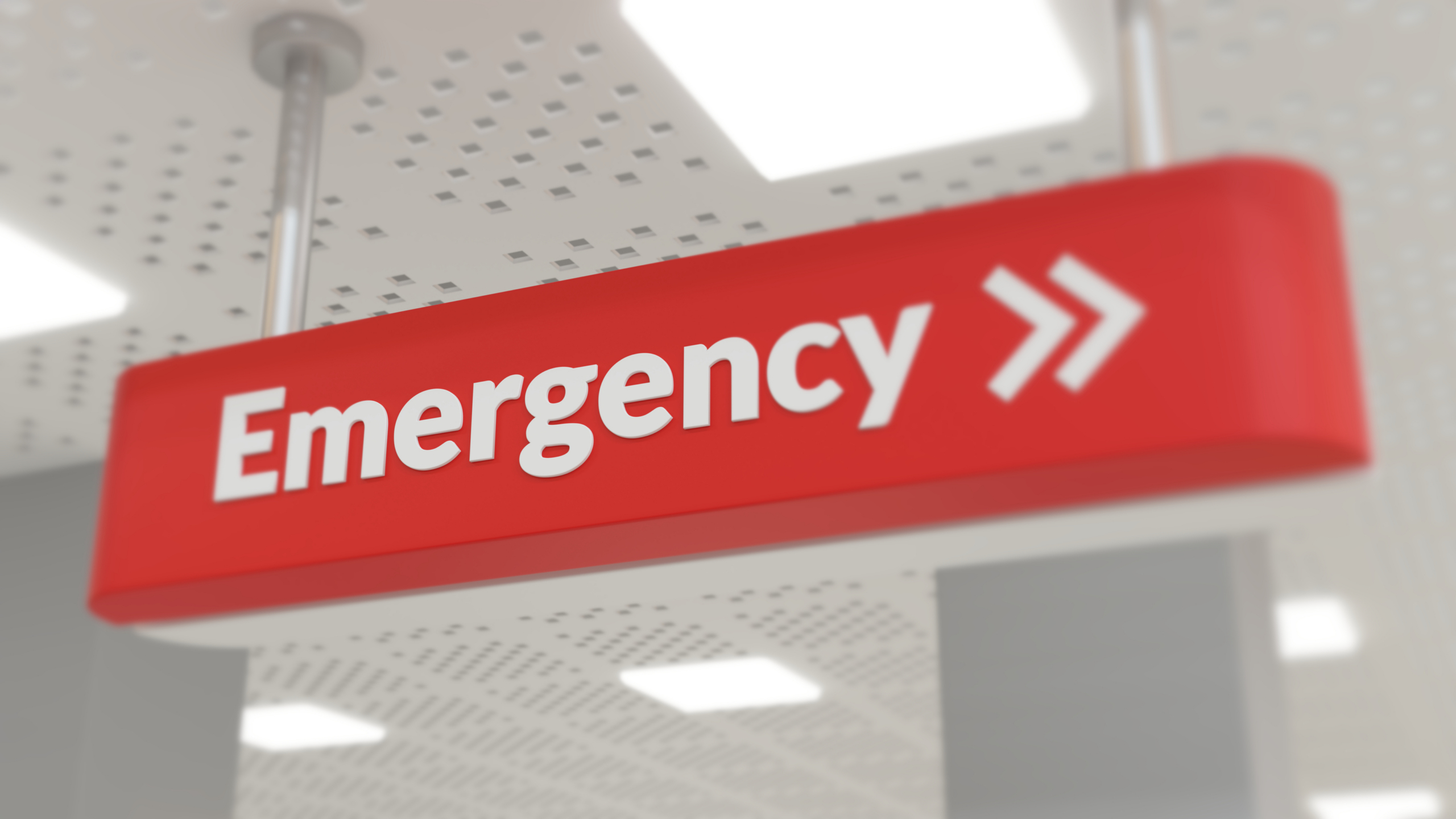 A red ceiling-mounted sign with white text reads “Emergency” followed by two right-pointing arrows, indicating the direction to the emergency department. The ceiling is white with rectangular lights and a dotted pattern.