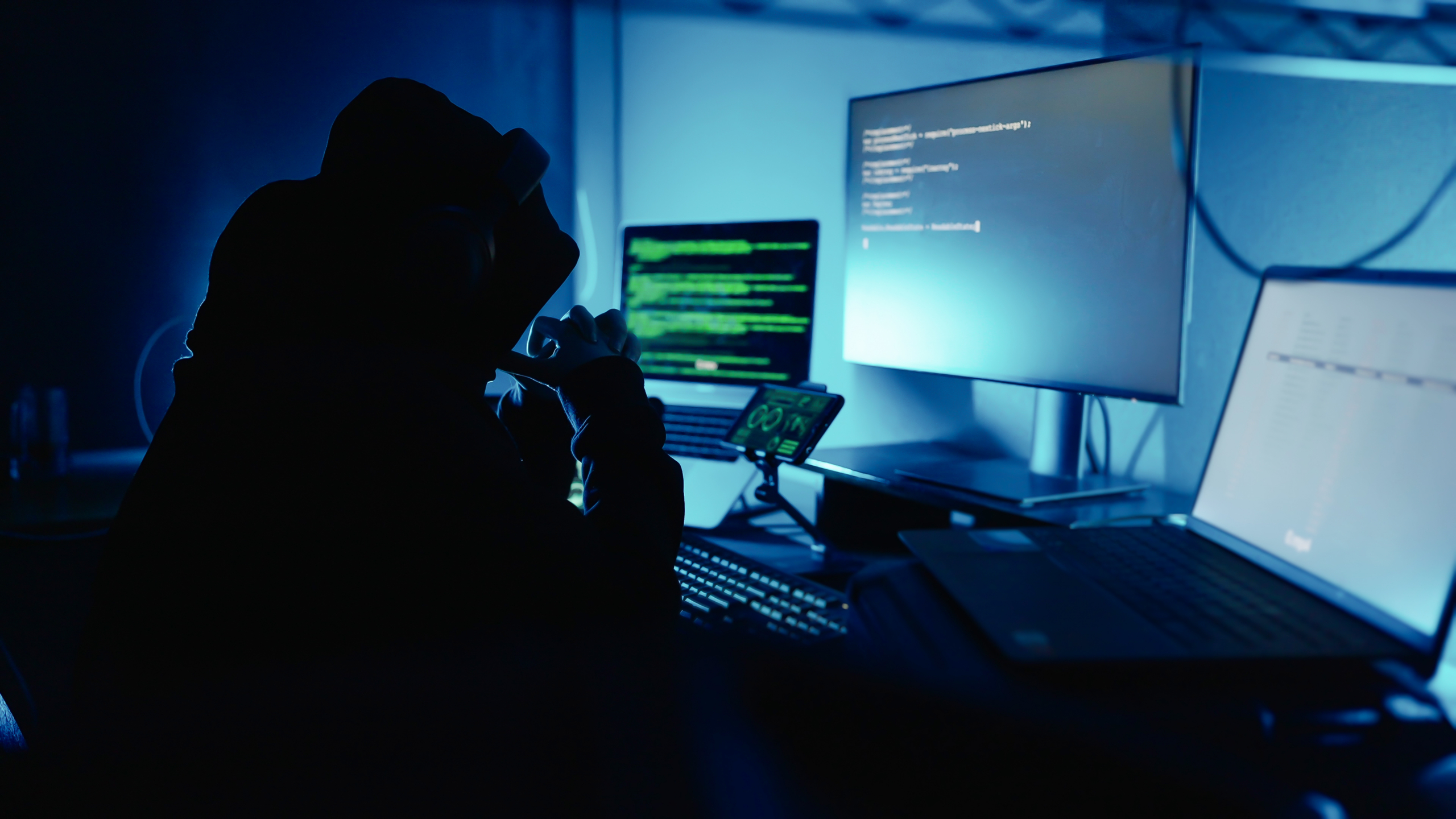 A person in a hoodie sits in a dark room, silhouetted against computer screens displaying code and data, suggesting hacking or cybersecurity activity.