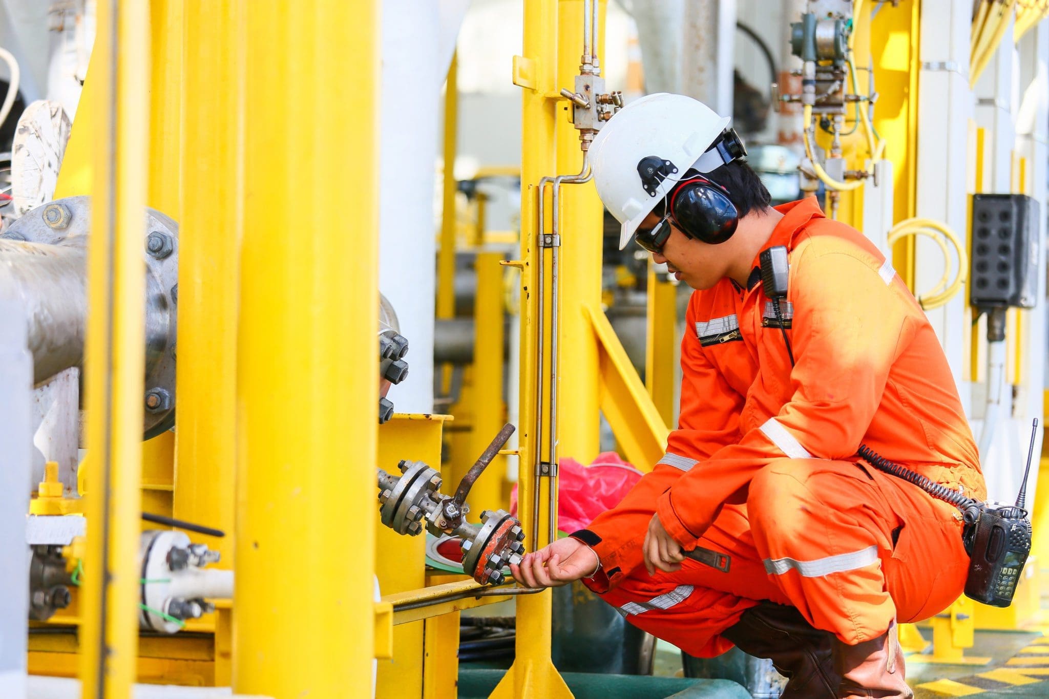 An engineer in orange safety clothing, helmet, and ear protection inspects equipment and valve controls at an industrial facility with yellow metal structures.