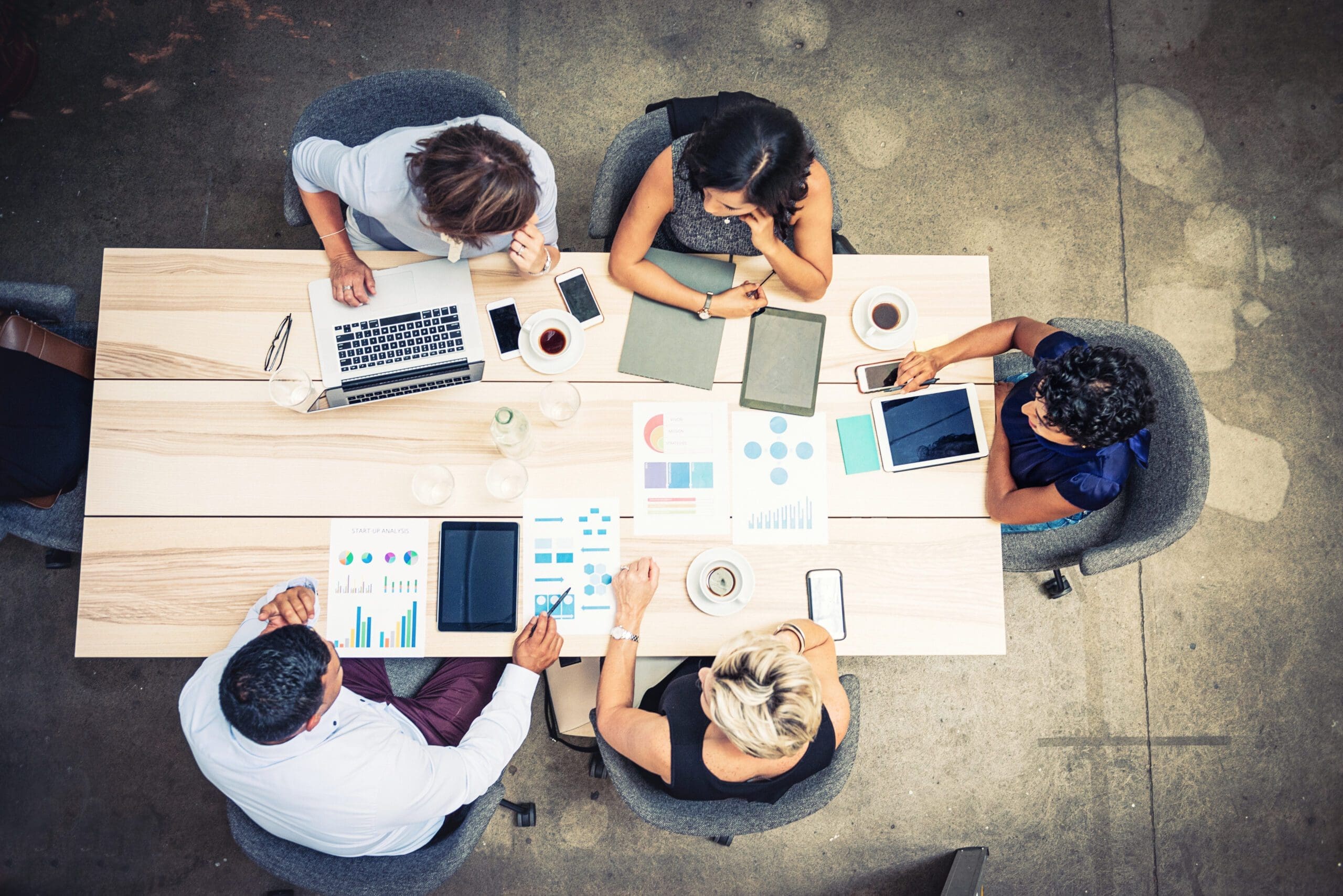 Five people sit around a table with charts, laptops, tablets, and coffee cups, engaged in a business meeting. The image is taken from above, showing a collaborative work environment.