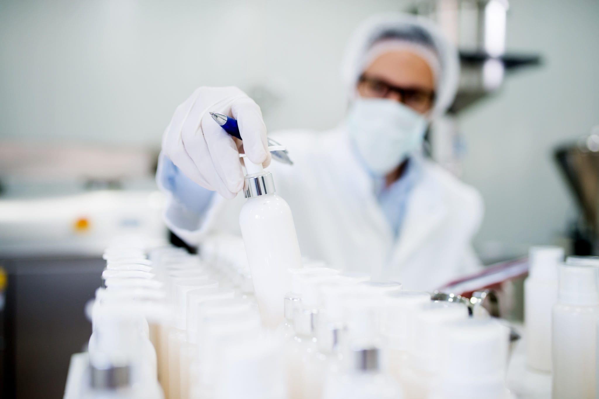 A scientist wearing a lab coat, gloves, hairnet, and mask examines a white plastic bottle among many similar bottles in a laboratory setting.