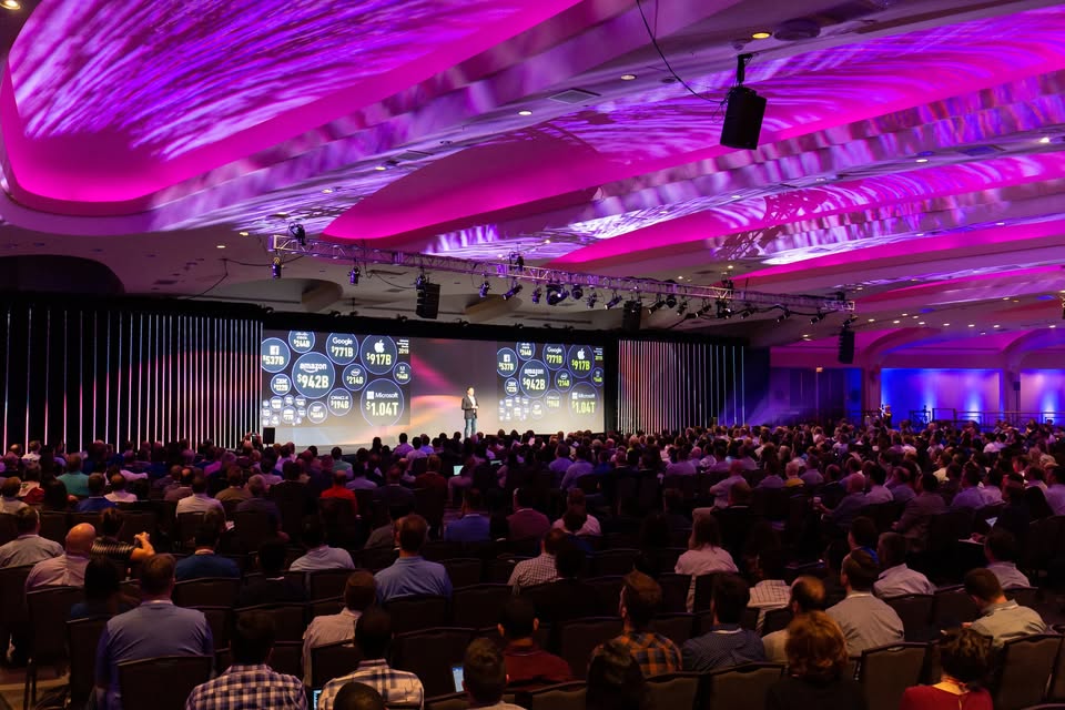 A large audience sits facing a brightly lit stage with a presenter speaking in front of colorful screens under purple ceiling lights at a conference or seminar.