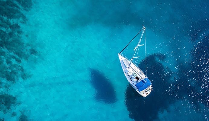 A white sailboat floats on calm, clear blue water, casting a shadow below. The sea appears turquoise and deepens in color, with sunlight reflecting off the surface. The scene is peaceful and serene.