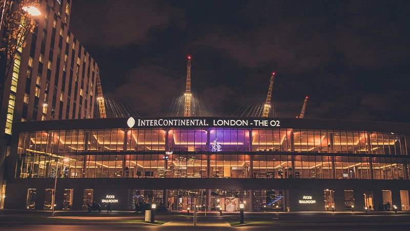 Night view of the InterContinental London - The O2 hotel, with large glass windows illuminated from inside. The O2 arena’s distinctive yellow masts are visible in the background against a dark sky.