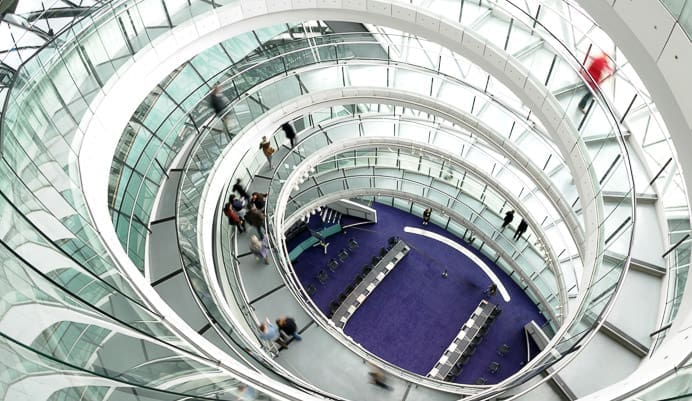 A top-down view of a modern building interior with a spiral staircase and glass railings. People are walking along the curved walkways, and there is a purple carpeted area with desks and chairs on the ground floor.