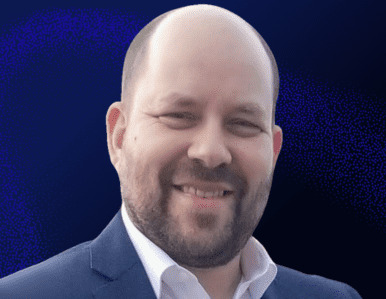 A smiling man with a beard, wearing a white shirt and a dark suit jacket, stands against a dark blue background.
