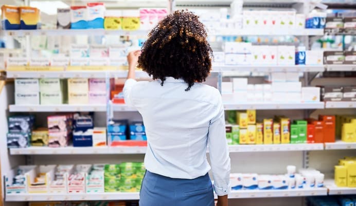A person with curly hair, seen from behind, stands in a pharmacy aisle reaching for a product on a shelf filled with various medicines and health items.