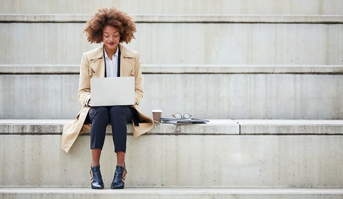 A woman sits alone on outdoor concrete steps, working on a laptop. She is wearing a tan trench coat, black pants, and ankle boots. A coffee cup, notebook, and glasses are placed on the step beside her.