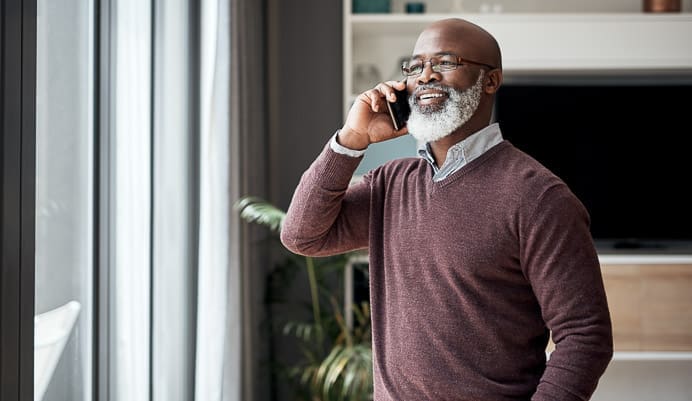 Smiling man with a gray beard and glasses stands indoors by a window, holding a smartphone to his ear while talking on the phone. He is wearing a maroon sweater over a collared shirt.