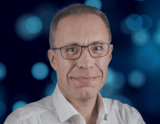 A middle-aged man with short hair and glasses, wearing a white shirt, smiles slightly at the camera. The background features blue bokeh lights.