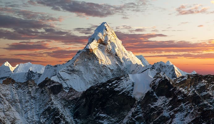 Snow-capped mountain peak illuminated by warm sunlight with dramatic clouds and an orange-pink sky in the background. Rugged, dark foreground ridges contrast with the bright summit.