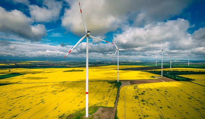 Several large wind turbines, serving as key elements of Identity for Critical Infrastructure, stand in a field of bright yellow flowers under a partly cloudy sky, with distant green fields and hills stretching to the horizon.