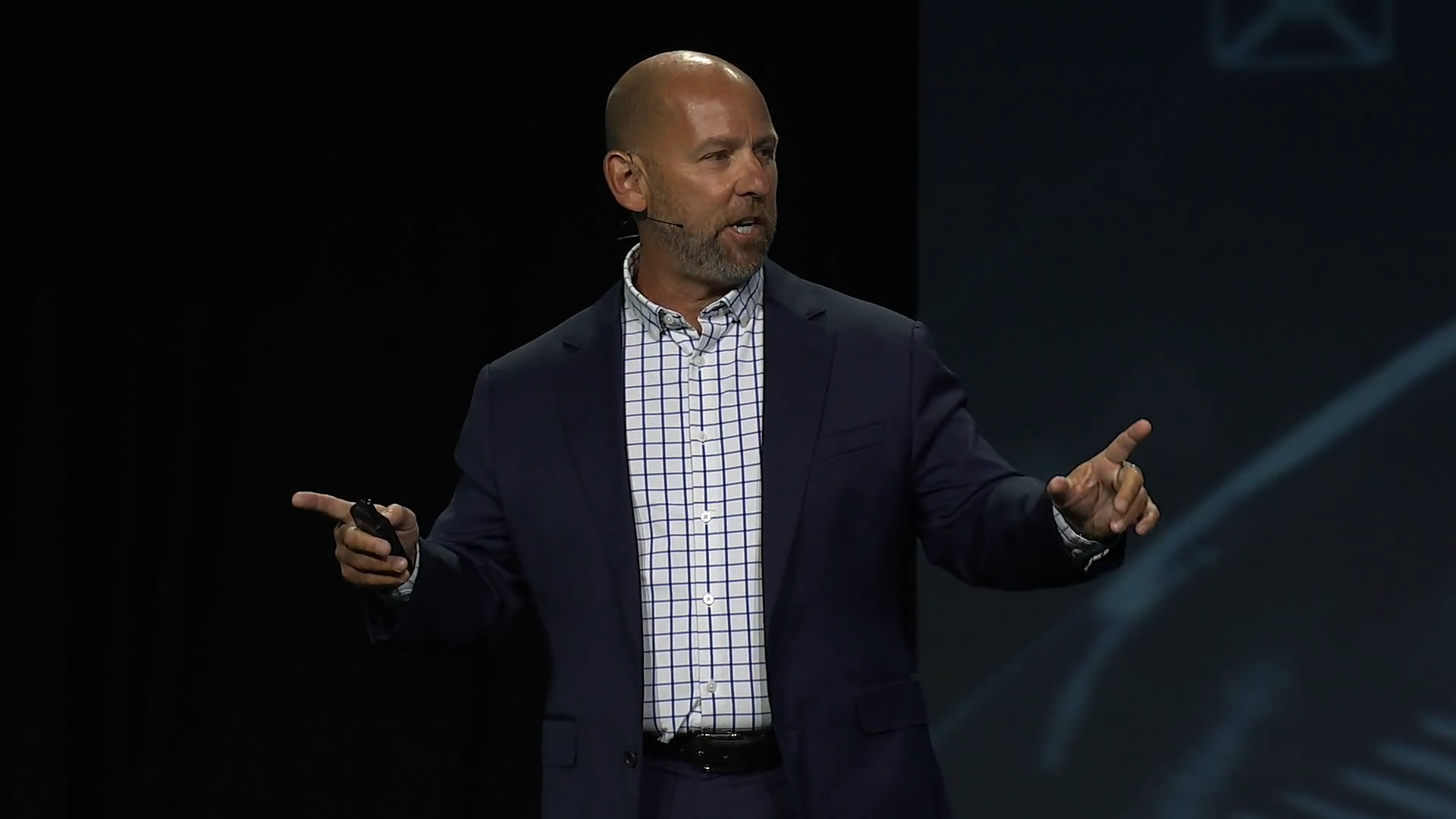 A bald man in a navy suit and checked shirt speaks on stage with a headset microphone, pointing with both hands against a dark background.