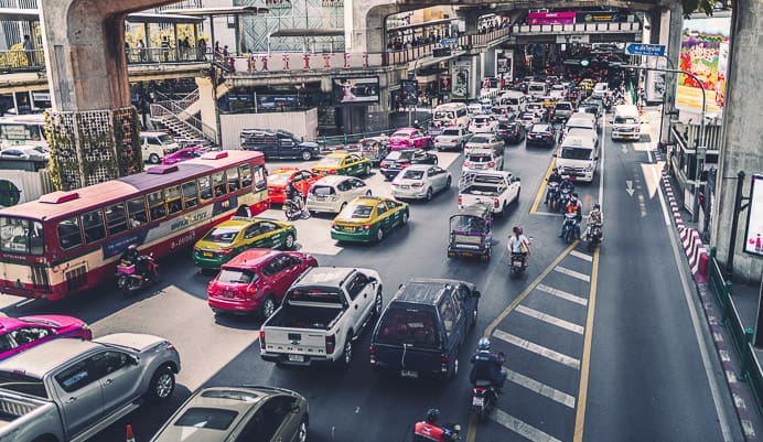 A busy urban street filled with cars, taxis, buses, and motorcycles, moving in multiple lanes beneath elevated trains and walkways on a sunny day.