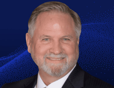 Smiling middle-aged man with gray hair and a beard, wearing a dark suit and white shirt, posed against a blue background with a dotted pattern.
